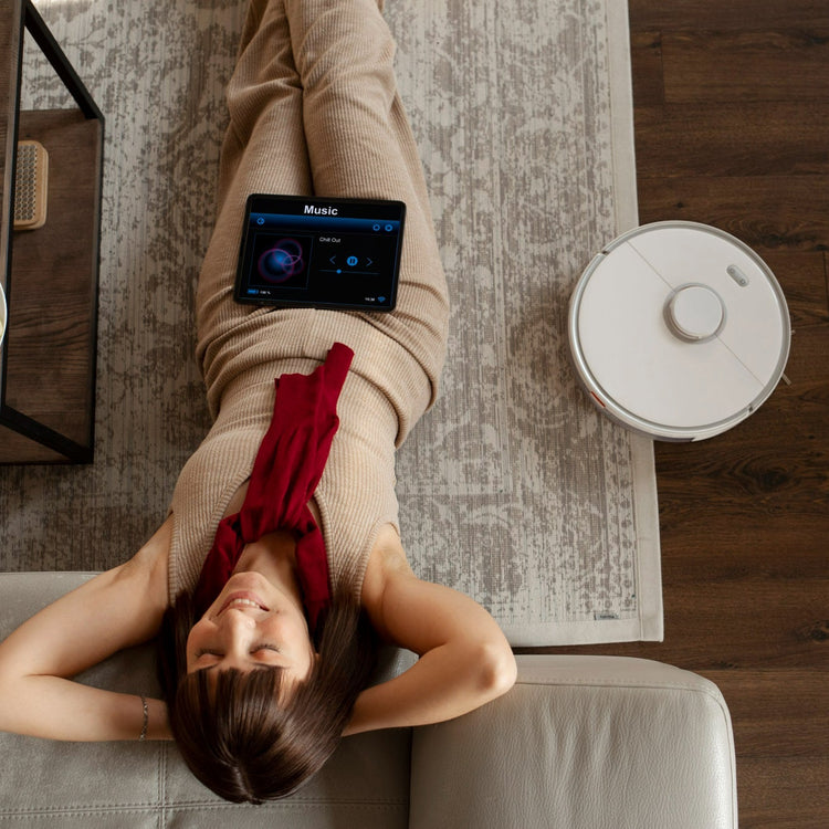 Person lying on a couch with a tablet and a robot vacuum cleaner in a room.